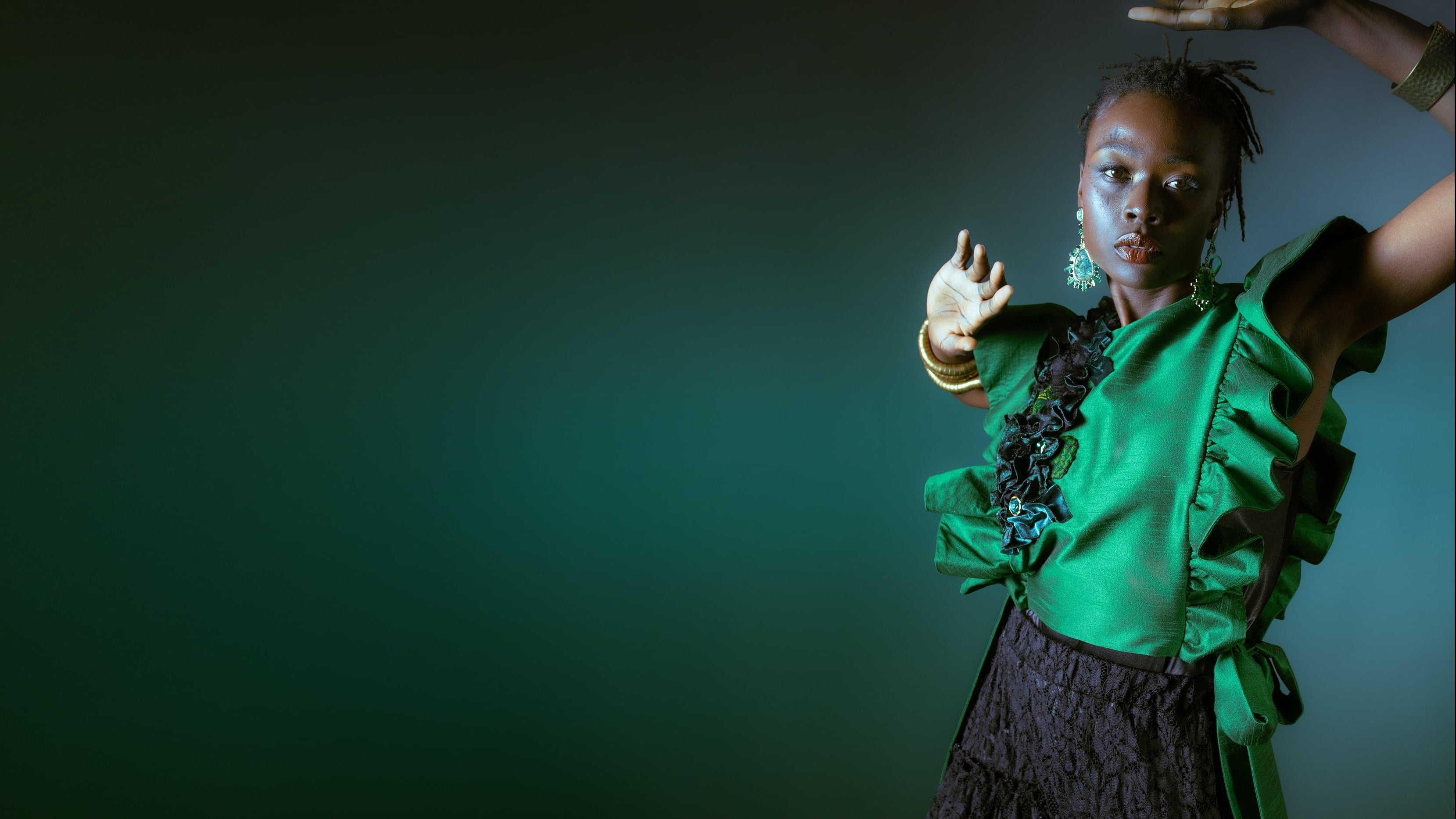 A model wearing a vibrant green ruffled blouse and a dark textured skirt poses against a dark green studio background with dramatic lighting.